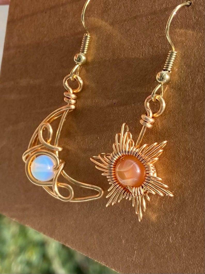 May include: Gold wire wrapped earrings with a crescent moon and sun design. The moon earring features a white opal stone and the sun earring features an orange carnelian stone.