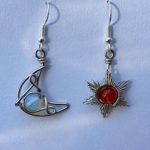 May include: A pair of silver earrings with a crescent moon and a star design. The moon earring features a white opal stone and the star earring has a red carnelian stone.