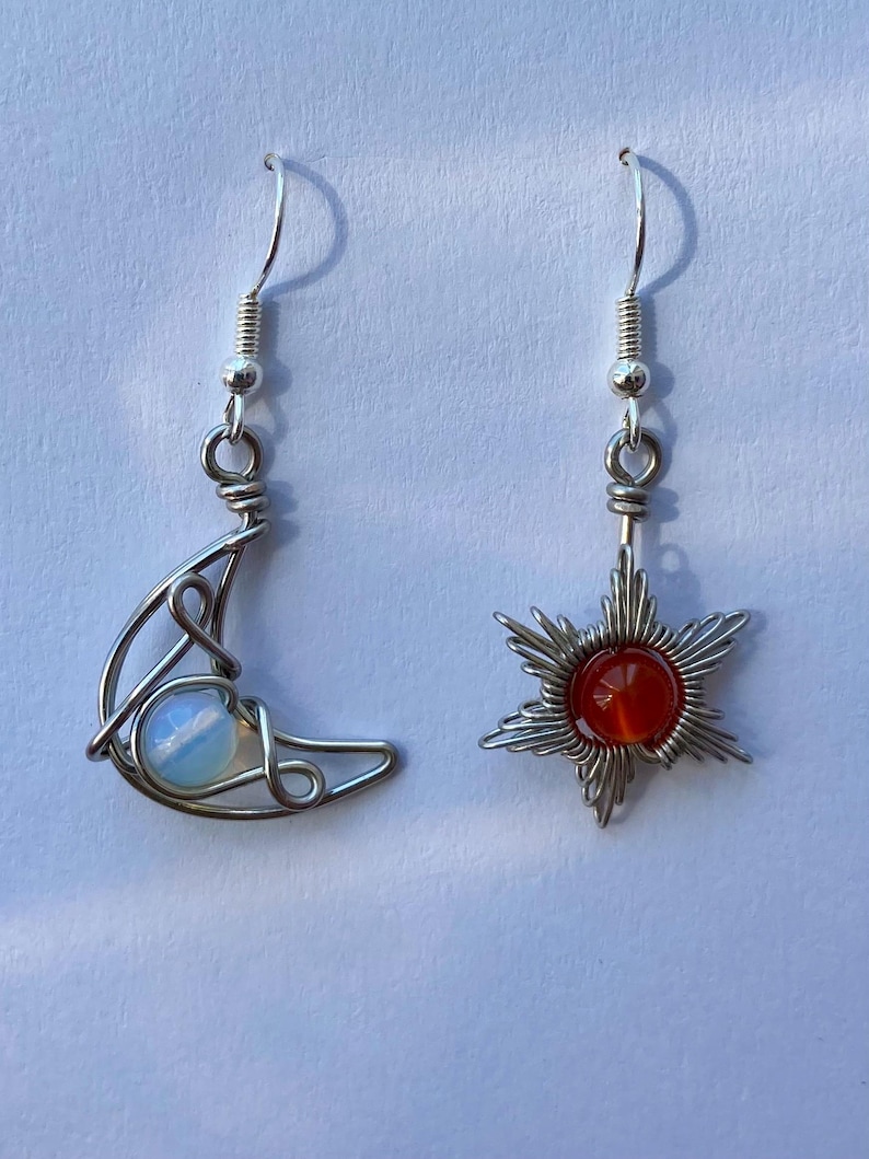 May include: A pair of silver earrings with a crescent moon and a star design. The moon earring features a white opal stone and the star earring has a red carnelian stone.