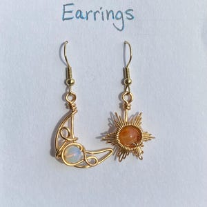 May include: Gold wire earrings with a crescent moon and a sun design. The moon earring has a white opal stone and the sun earring has an orange carnelian stone. The earrings are labelled "Sun & Moon Earrings".