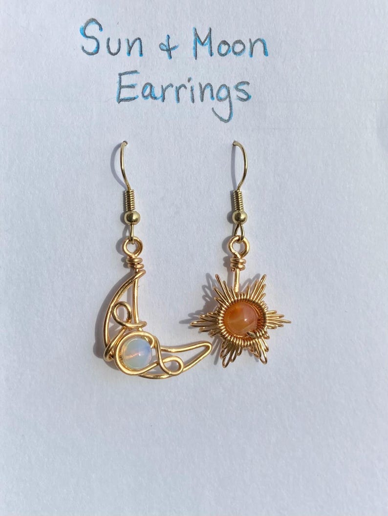 May include: Gold wire earrings with a crescent moon and a sun design. The moon earring has a white opal stone and the sun earring has an orange carnelian stone. The earrings are labelled "Sun & Moon Earrings".