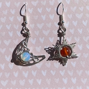 May include: A pair of silver earrings with a crescent moon and a star design. The moon earring features a light blue opal stone, while the star earring has a red carnelian stone.