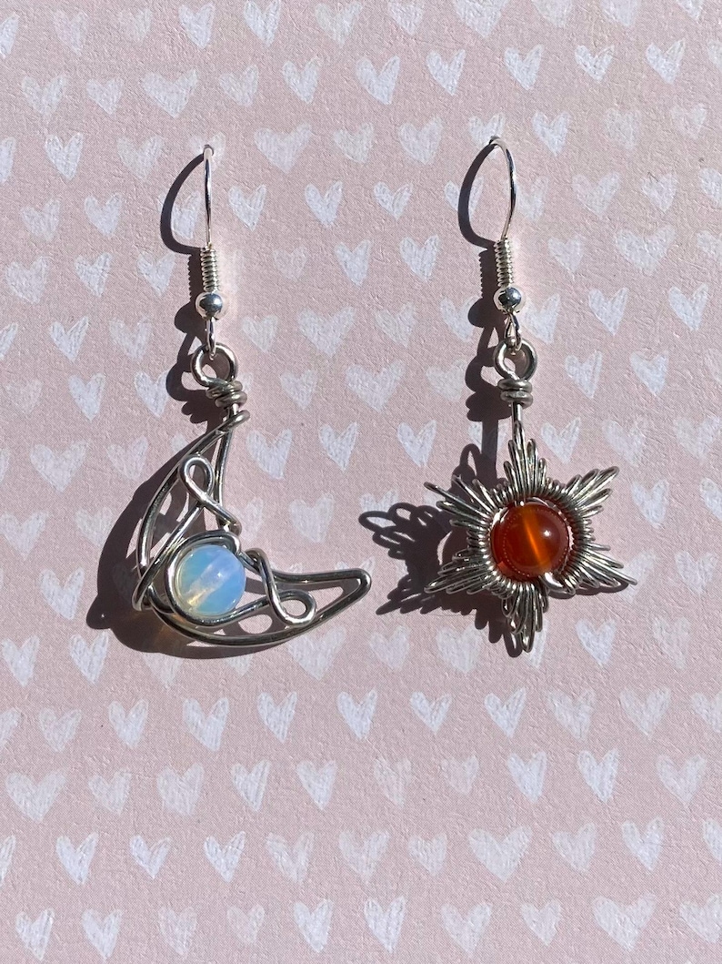 May include: A pair of silver earrings with a crescent moon and a star design. The moon earring features a light blue opal stone, while the star earring has a red carnelian stone.