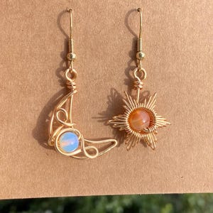 May include: A pair of gold wire earrings with a moon and star design. The moon earring features a light blue opal stone, while the star earring has an orange carnelian stone.