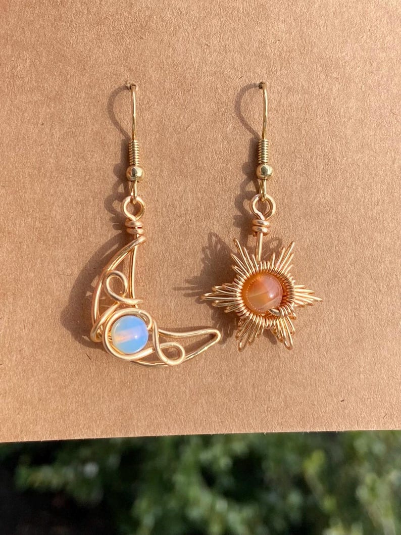 May include: A pair of gold wire earrings with a moon and star design. The moon earring features a light blue opal stone, while the star earring has an orange carnelian stone.