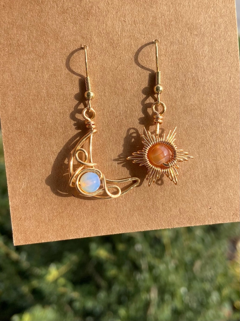 May include: A pair of gold wire earrings with a crescent moon and a sun design. The moon earring has a white opal stone and the sun earring has an orange stone.