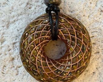 May include: A handcrafted pendant necklace featuring a circular, textured pendant with a copper wire grid design. The pendant has a central hole and is suspended from a black cord. The pendant is a mix of green, brown, and gold tones.