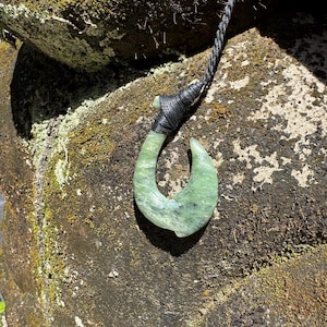 May include: A green jade hook pendant with a black cord. The hook is carved from a light green stone and has a curved shape. The cord is black and appears to be braided. The pendant is displayed against a mossy rock background.