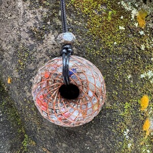 May include: A clear, round pendant necklace with a black cord. The pendant features a spiral design of copper wire and colorful stones. The necklace is displayed against a mossy, textured background.