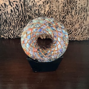 Può includere: Una scultura decorativa in vetro con forma circolare e apertura centrale. La scultura presenta un motivo a mosaico di pezzi di vetro blu, rossi e arancioni, racchiusi in una griglia di filo di rame. Poggia su un piccolo supporto nero.