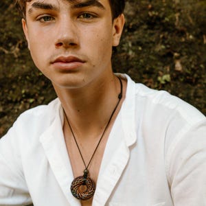 May include: A circular pendant necklace with a woven design in shades of brown, orange, and green. The pendant hangs from a black cord. The person is wearing a white button-down shirt.