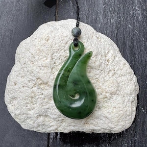 May include: A green jade pendant in a hook shape, suspended from a black cord with a small bead. The pendant has a polished, glossy finish and is displayed on a textured, off-white stone. The background is a dark, slate-like surface.
