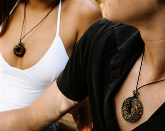 May include: Two people wearing pendant necklaces. The necklaces have a circular design with green and gold wire wrapped around a dark centre. The pendants hang from black cords. One person wears a white top, and the other wears a black shirt.