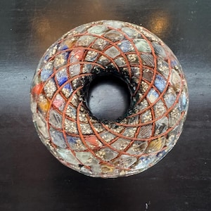 May include: A donut-shaped orgone energy device. The device is made of clear, multi-colored, and square-cut stones, encased in a copper wire grid. The center hole is black, and the overall appearance is textured.