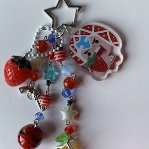 Puede incluir: Un llavero con un cierre metálico en forma de estrella, un dije de acrílico transparente de un personaje con sombrero de patrón rojo y blanco comiendo una fresa, y varias cuentas colgantes que incluyen una fresa roja, un revólver plateado y estrellas.