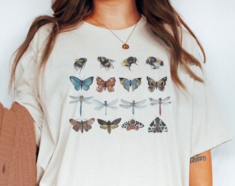 May include: A cream-coloured short-sleeve t-shirt featuring a graphic print of various insects arranged in four rows. The design includes illustrations of bees, butterflies, dragonflies, and moths in shades of blue, brown, yellow, and black.