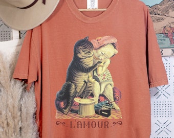 May include: Rust-colored t-shirt featuring a vintage illustration of two cats, one in a hat and the other holding a fan. The text "L'AMOUR" is below the image. The shirt is hanging on a wooden hanger.