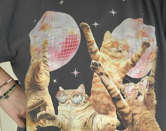 May include: A black t-shirt with a graphic of three cats wearing sunglasses and holding a disco ball. The cats are standing in front of a record player with two records on it. The background is a dark blue with stars.
