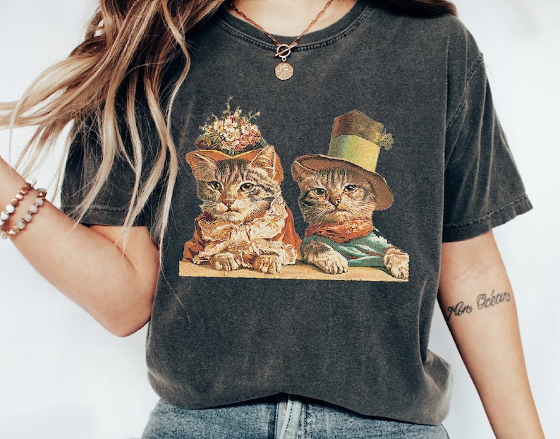 May include: A dark gray t-shirt with a vintage illustration of two cats wearing hats. The cat on the left is wearing a brown hat with flowers and the cat on the right is wearing a green top hat. The text "Mon Oc&eacute;an" is tattooed on the wearer's arm.