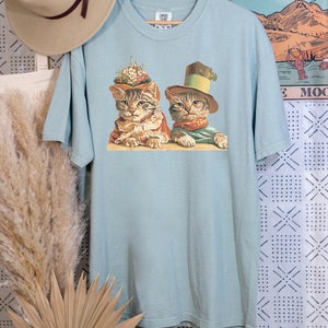 May include: A light blue t-shirt with a vintage illustration of two cats wearing hats. The cats are sitting side-by-side and looking at the viewer. The illustration is in a muted color palette with shades of brown, green, and white.