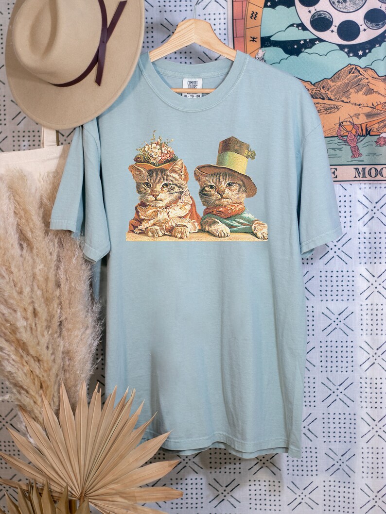 May include: A light blue t-shirt with a vintage illustration of two cats wearing hats. The cats are sitting side-by-side and looking at the viewer. The illustration is in a muted color palette with shades of brown, green, and white.