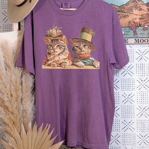 May include: Purple t-shirt with a vintage illustration of two cats wearing hats. The cat on the left is wearing a flower crown and the cat on the right is wearing a top hat.