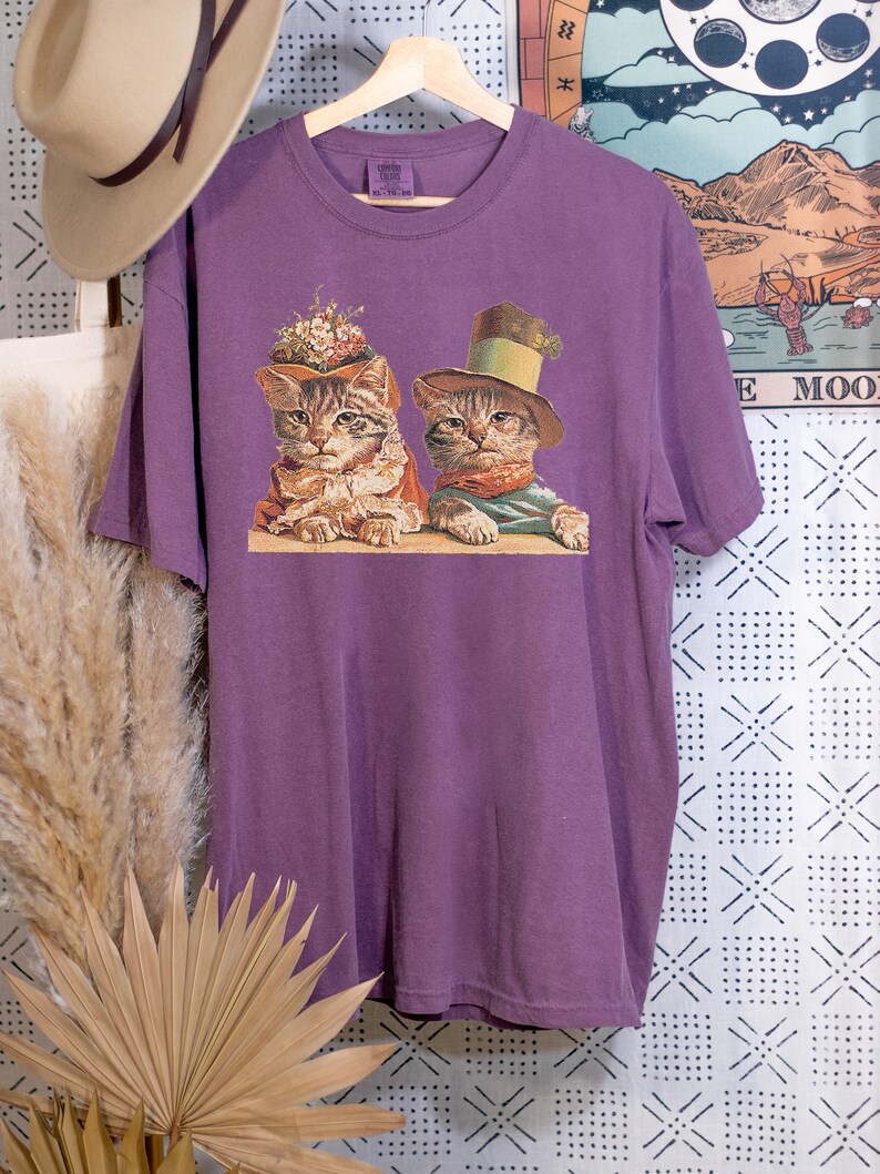 May include: Purple t-shirt with a vintage illustration of two cats wearing hats. The cat on the left is wearing a flower crown and the cat on the right is wearing a top hat.