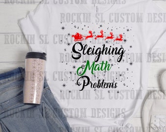Op de afbeelding: Wit T-shirt met een kerstthema met een rood slee met rendieren die door een besneeuwde lucht vliegen. De tekst "Sleighing Math Problems" is geschreven in zwarte en groene letters.