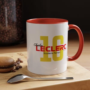 Könnte beinhalten: Weiße Keramiktasse mit rotem Rand und Henkel. Die Tasse zeigt eine gelbe Nummer "16" mit dem Text "Charles Leclerc Scuderia Ferrari" in Rot und Gelb.