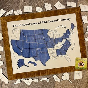 May include: A wooden map of the United States with a brown frame. The map is divided into states, each with a blue background and white lettering. The states are arranged in a traditional map format. The title "The Adventures of The Everett Family" is written at the top of the map. There are individual state pieces surrounding the map, each with a white background and black lettering.