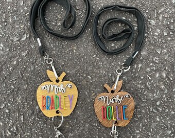 May include: Two wooden apple-shaped lanyards with keychains. One lanyard says "Mrs. Bradley" with rainbow lettering and the other says "Nurse Dollie" with rainbow lettering.