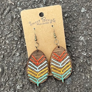 May include: A pair of wooden earrings with a leaf design. The leaves are painted with a chevron pattern in shades of orange, yellow, white, and blue. The earrings are attached to gold-toned hooks.