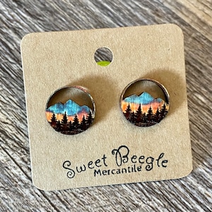 May include: A pair of wooden earrings with a mountain scene and sunset sky. The earrings are round and have a brown wood finish. The text "Sweet Beagle Mercantile" is printed on the card.