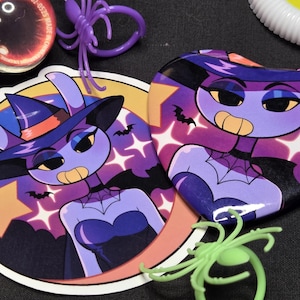 May include: A round sticker and a heart-shaped pin featuring a purple-skinned character wearing a witch hat and a black dress. The background shows a dark sky with stars and bats. A purple plastic spider ring and a light green plastic spider ring.