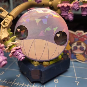 May include: A whimsical, round, purple figure with a holographic surface and a wide, toothy grin. The figure has large eyes with heart-shaped pupils and sits atop a blue and green base. Decorative elements surround the figure.