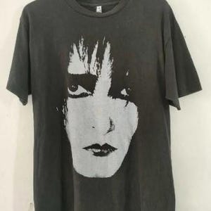 May include: A dark gray t-shirt featuring a large, stylized black and white portrait of a person's face. The face has prominent eyes and dark lips. The t-shirt is hanging on a wooden hanger.