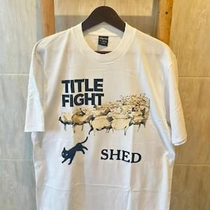 May include: White t-shirt with a graphic design featuring the words "TITLE FIGHT" above a flock of sheep and the word "SHED" below. A black cat is jumping in front of the sheep. The shirt is hanging on a wooden hanger.