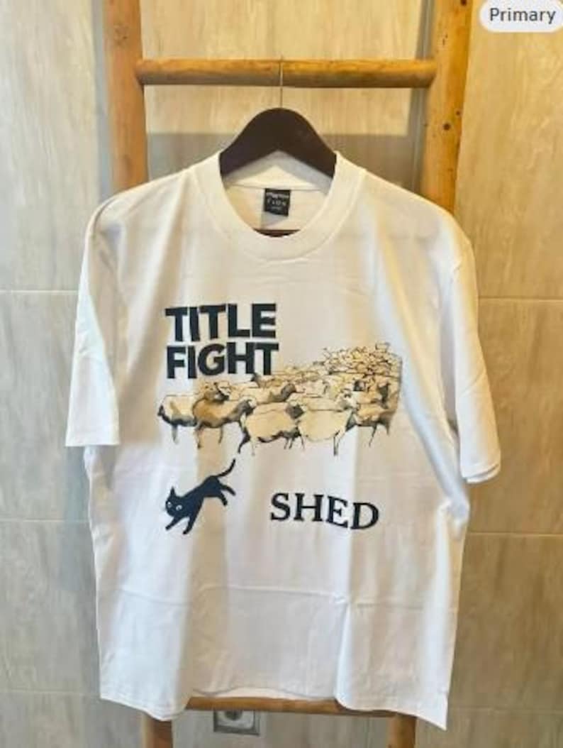 May include: White t-shirt with a graphic design featuring the words "TITLE FIGHT" above a flock of sheep and the word "SHED" below. A black cat is jumping in front of the sheep. The shirt is hanging on a wooden hanger.