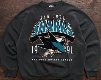 May include: Dark gray sweatshirt featuring the San Jose Sharks logo. The logo includes a teal and black shark, the word "SHARKS" in teal and white, and the years "19" and "91". The words "NATIONAL HOCKEY LEAGUE" are printed below the logo.