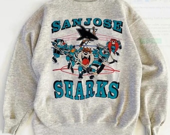 May include: A light gray sweatshirt featuring the words "SAN JOSE SHARKS" in teal block letters. The graphic depicts cartoon characters playing hockey, with a black shark logo above. The design includes red hockey lines.