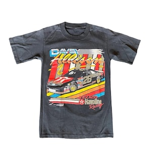 May include: Dark gray t-shirt featuring a colorful graphic of a race car with the number 28. The shirt displays the text "Davey Allison" and "Texaco Havoline Racing" in a vintage style.