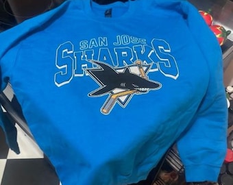 May include: A bright blue San Jose Sharks sweatshirt. The sweatshirt features the team name and logo in white and black, with a shark graphic. The long-sleeved crew neck top is made of a soft material.