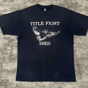 Pode incluir: Camiseta preta com as palavras "TITLE FIGHT" acima de um gr&aacute;fico branco de uma coruja com as asas abertas e a palavra "SHED" abaixo. A camiseta &eacute; de gola redonda.