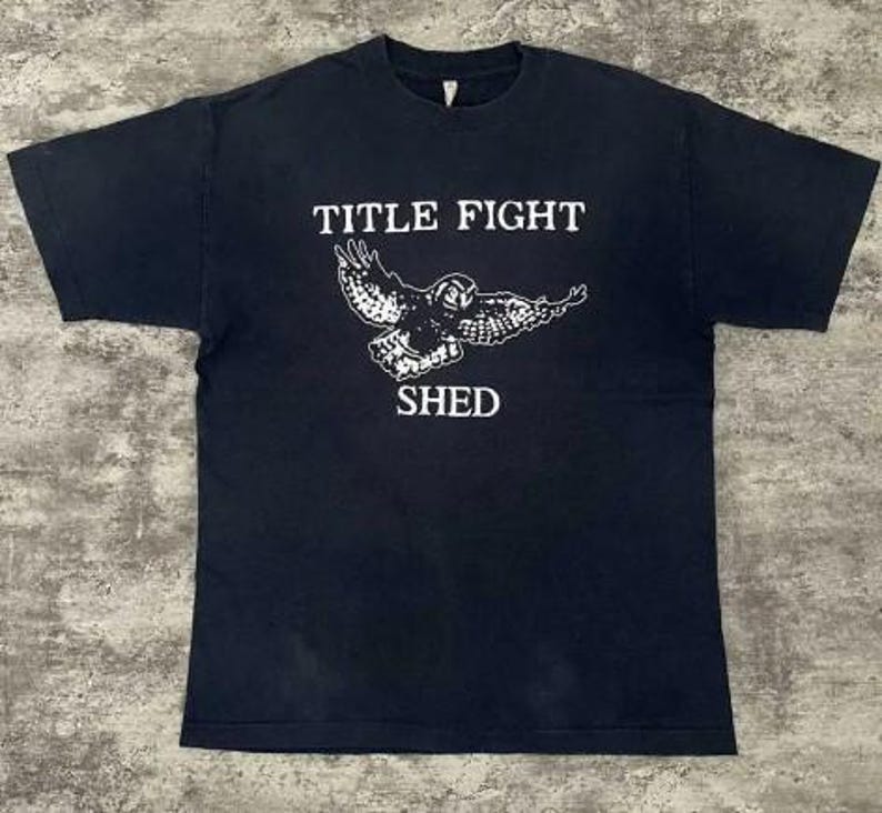 Pode incluir: Camiseta preta com as palavras "TITLE FIGHT" acima de um gr&aacute;fico branco de uma coruja com as asas abertas e a palavra "SHED" abaixo. A camiseta &eacute; de gola redonda.
