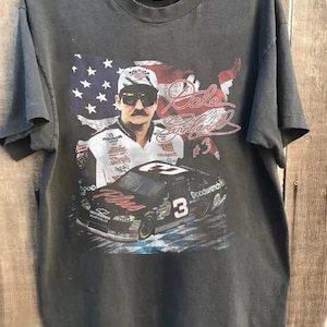 K&ouml;nnte beinhalten: Dunkelgraues T-Shirt mit einer Grafik von Dale Earnhardt, einem ber&uuml;hmten NASCAR-Fahrer, mit seinem Bild, der Nummer 3 und einem Rennwagen. Die amerikanische Flagge ist im Hintergrund. Das T-Shirt h&auml;ngt an einem Holzb&uuml;gel.