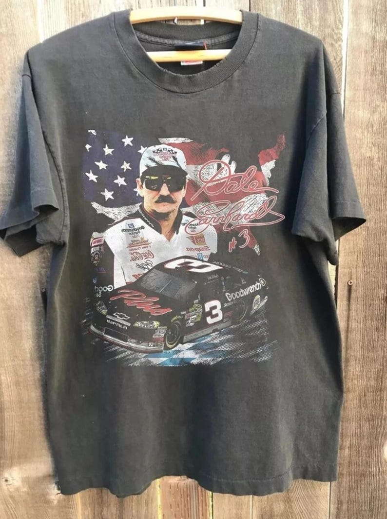 K&ouml;nnte beinhalten: Dunkelgraues T-Shirt mit einer Grafik von Dale Earnhardt, einem ber&uuml;hmten NASCAR-Fahrer, mit seinem Bild, der Nummer 3 und einem Rennwagen. Die amerikanische Flagge ist im Hintergrund. Das T-Shirt h&auml;ngt an einem Holzb&uuml;gel.