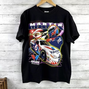 May include: Black t-shirt featuring a graphic of a race car with the number 6 and the text "Valvoline Racing." The design includes a driver in a helmet and the name "MARTIN". The shirt is hanging on a wooden hanger.