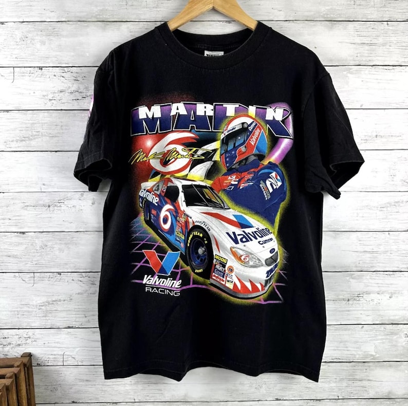 May include: Black t-shirt featuring a graphic of a race car with the number 6 and the text "Valvoline Racing." The design includes a driver in a helmet and the name "MARTIN". The shirt is hanging on a wooden hanger.
