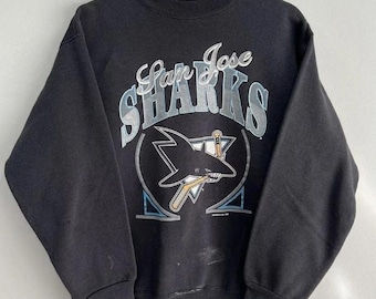 May include: Black San Jose Sharks sweatshirt with a crew neck. The front features the team logo with the words "San Jose Sharks" in a stylized font above the logo. The logo includes a shark with a hockey stick.