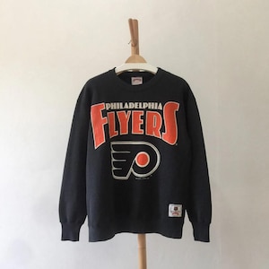 Może przedstawiać: Ciemnoszara bluza Philadelphia Flyers z pomarańczowym napisem i logo. Słowo "FLYERS" jest dużymi pomarańczowymi literami nad logo drużyny. Logo drużyny to stylizowane pomarańczowe koło w czarno-białym wzorze.